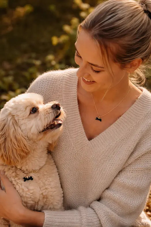 Woman in a beige sweater holding a white dog outdoors with greenery in the background